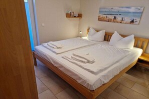 Un lit double avec du linge de lit et des serviettes blanches, une tête de lit en bois, une petite table de chevet avec une lampe, et un tableau sur le thème de la plage accroché au mur.