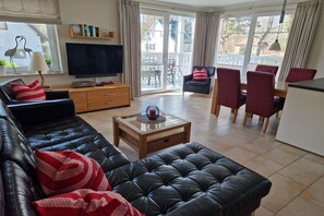 Ein Wohnzimmer mit schwarzem Ledersofa, einem Fernseher, einem Couchtisch und einem Essbereich mit roten Stühlen.