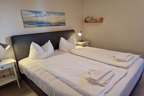 A double bed with white linens and pillows, a bedside table with a lamp, and a painting of a beach scene on the wall. - Holiday apartment with 2 bedrooms (Heringsdorf)