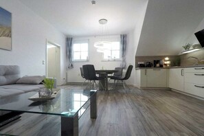 Ein zeitgemäßer Wohnraum mit einem Glastisch, einer Couch, einem Essbereich mit Stühlen und einer Küche mit Schränken.