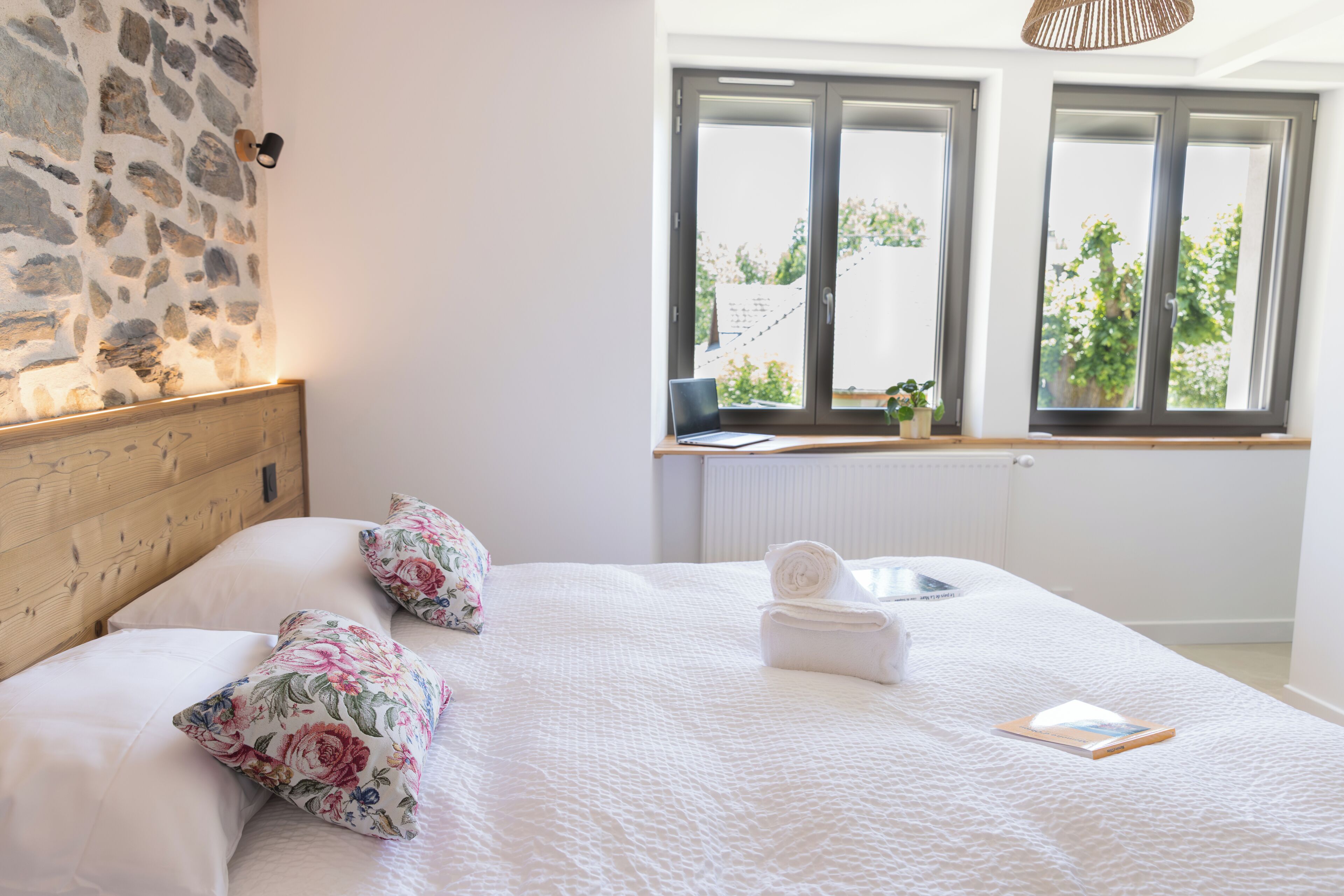 Ein Schlafzimmer mit Steinwand, einem Holz-Kopfteil, einem Bett mit weißer Bettwäsche, floralen Kissen, einem aufgerollten Handtuch und einem Buch.