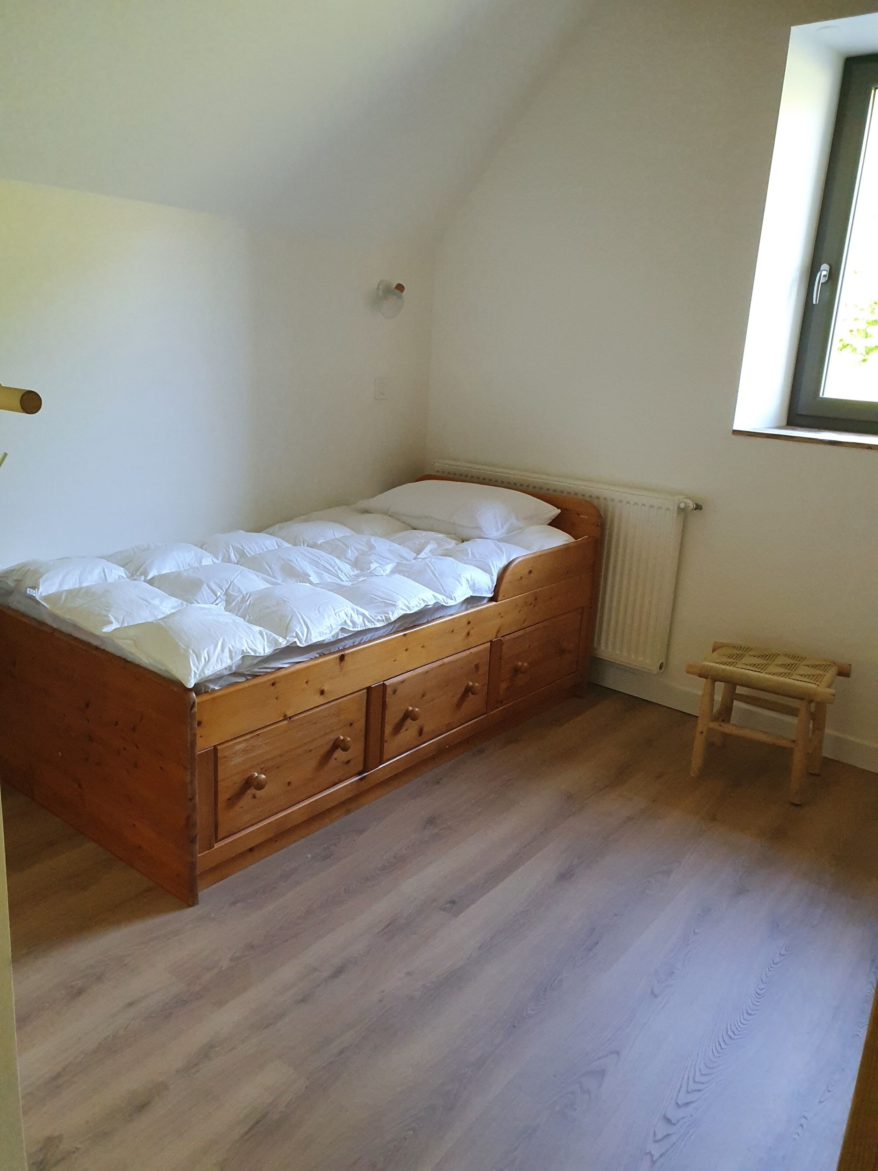 A small, single-bed room with a wooden bed frame and a small wooden stool.