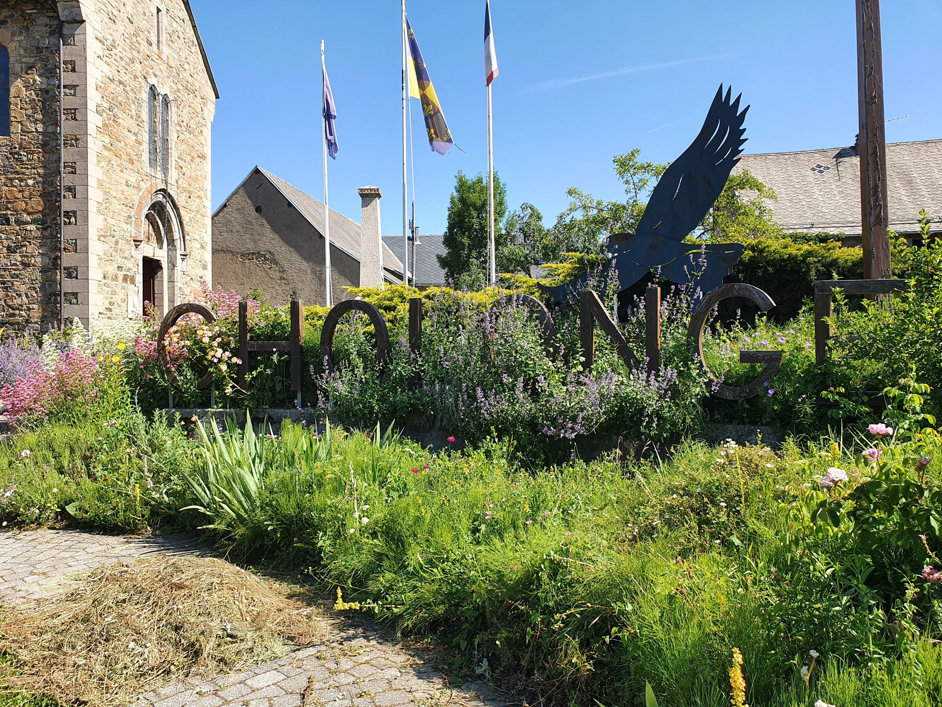 A garden with a variety of plants and flowers, a stone building with a chimney, and flags on flagpoles.