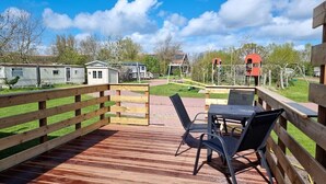 A wooden deck with a table and chairs, surrounded by a wooden fence, with a view of mobile homes and a playground in the background. - Beautiful pylon OK21 (Oostkapelle)