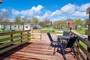 Eine Holzterrasse mit Tisch und Stühlen, umgeben von einem Holzzaun, mit Blick auf Mobilheime und einen Spielplatz im Hintergrund.