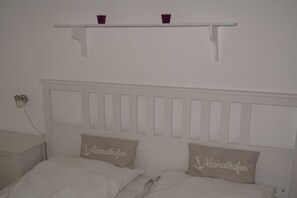 A bedroom with a white headboard, two pillows with text, a bedside table, and a shelf above the bed. - Strandnest Sierksdorf (Sierksdorf)