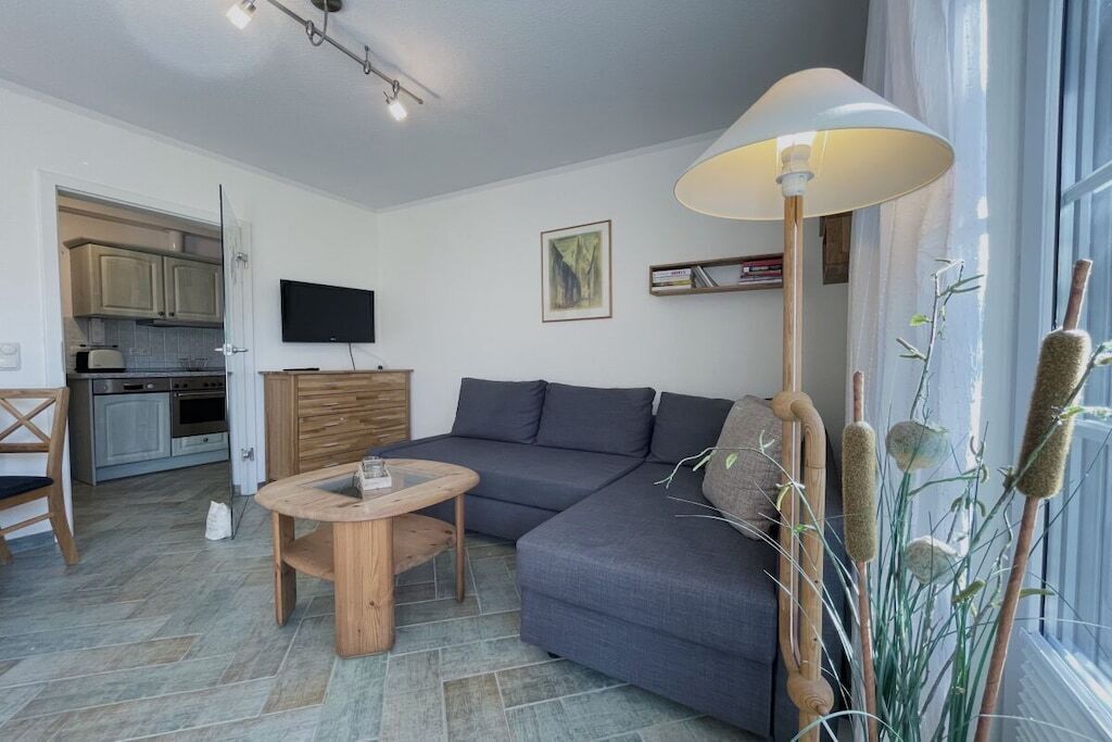 Ein modernes Wohnzimmer mit grauem Sofa, einem Holztisch, einem an der Wand montierten Fernseher und einem großen Fenster mit Tageslicht.