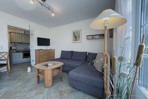 Ein modernes Wohnzimmer mit grauem Sofa, einem Holztisch, einem an der Wand montierten Fernseher und einem großen Fenster mit Tageslicht.