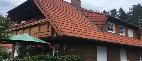 Una casa de dos pisos con techo de tejas rojas inclinadas, un balcón adornado con flores y una sombrilla verde.
