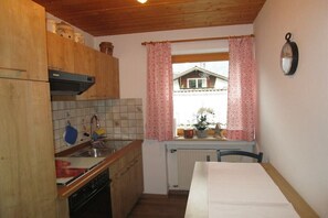 Cuina petita amb armaris de fusta, un salvasol de rajola, una finestra amb cortines roses i vista a una casa a l'exterior.