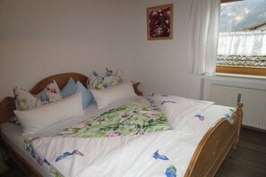 Ein Bett mit blumigem Bettbezug und vielen Kissen, ein Holz-Kopfteil, ein Fenster mit weißen Vorhängen und ein an der Wand hängendes Bild.