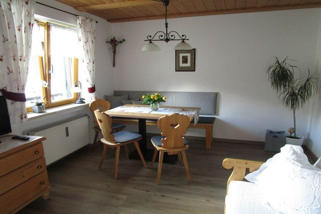 Ein Zimmer mit einem Holztisch, zwei Stühlen, einer Bank, einem Heizkörper, einem Fenster mit Vorhängen, einer Pflanze und einer Wandleuchte.