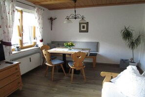 Een kamer met een houten eettafel, twee stoelen, een bankje, een radiator, een raam met gordijnen, een plant en een wandlamp.