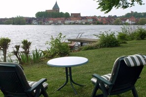 Ein Außenbereich mit Sitzgelegenheit, einem Tisch und zwei Stühlen, mit Blick auf ein Gewässer und Gebäude im Hintergrund.