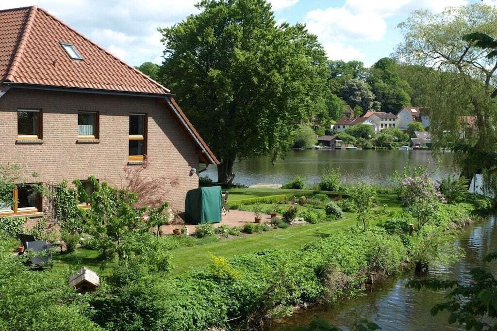 Ein Backsteinhaus mit Ziegeldach, ein Garten mit verschiedenen Pflanzen und ein Gewässer im Hintergrund.