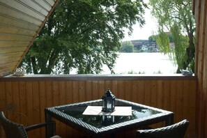 Eine Holzterrasse mit einem Glastisch und Korbstühlen, mit Blick auf ein Gewässer und Bäume.