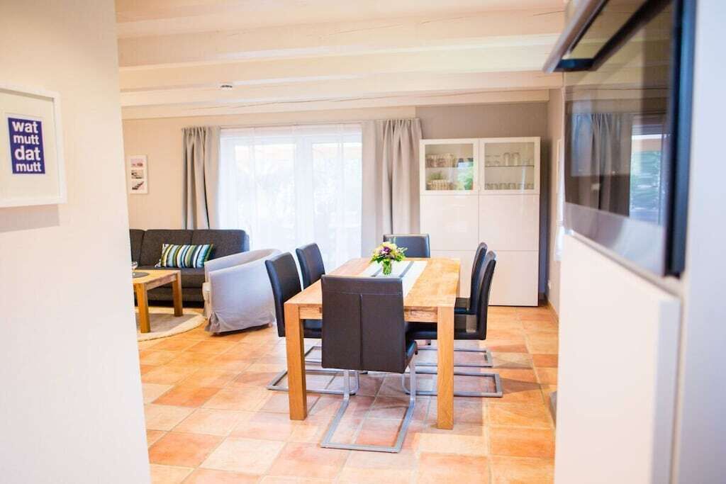 Ein Essbereich mit Holztisch, Stühlen und einer Blumenarragemment in der Mitte. Im Hintergrund befinden sich eine Couch und ein Schrank.