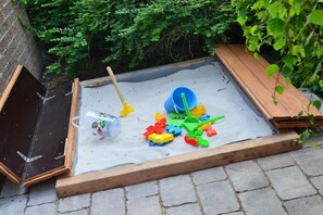 Ein Sandkasten mit Spielzeug und einer Schaufel, umgeben von einem Holzrahmen und Grünpflanzen.
