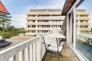 A balcony with white benches, a car parked outside, and a modern building with multiple balconies in the background. - Villa by the sea, holiday apartment 8 (St. Peter-Ording)