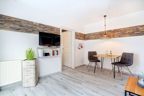 Ein modernes Wohnzimmer mit Fernseher, Bücherregal, Essbereich mit zwei Stühlen und einer Pendelleuchte.