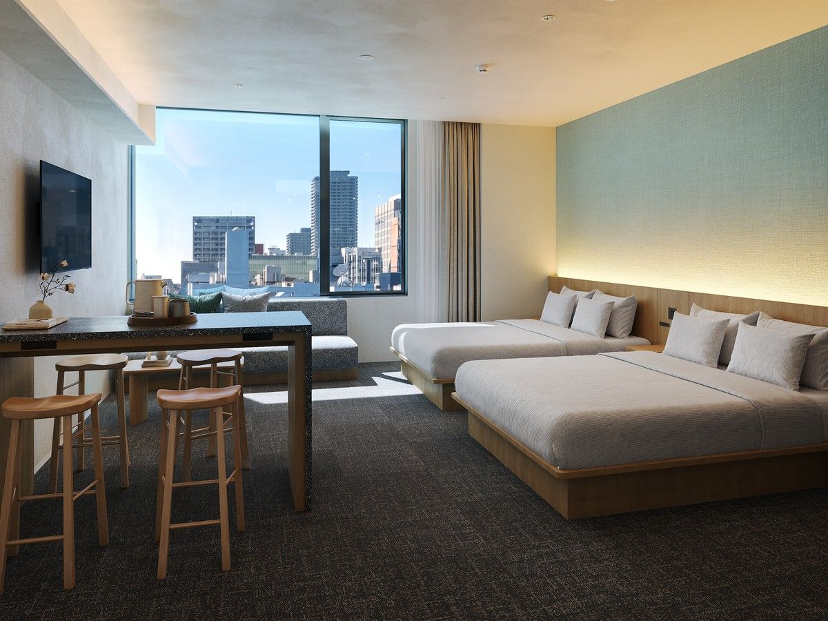 Habitación de hotel con un ventanal grande, un escritorio con silla, dos camas y un taburete.