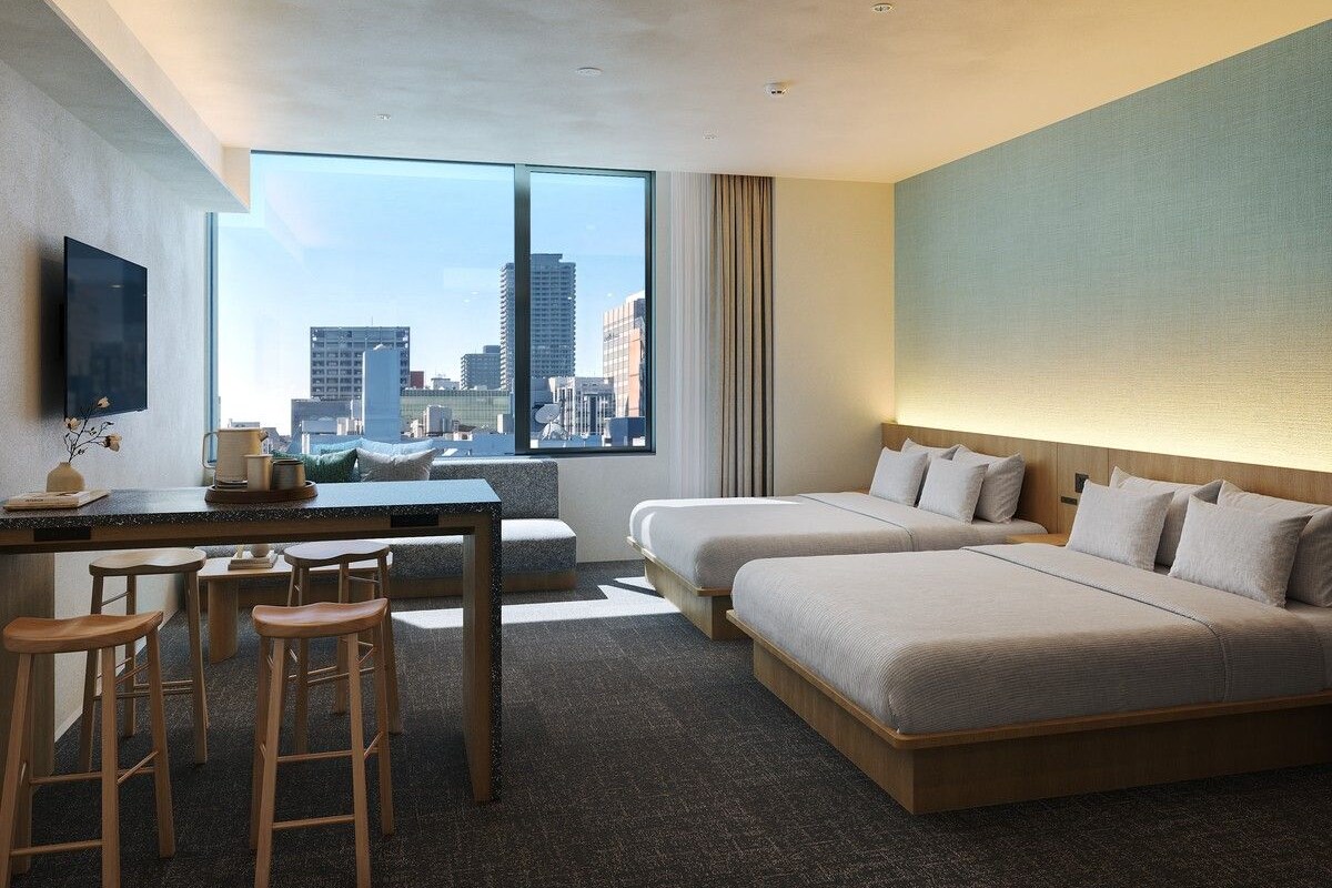 Habitación de hotel con un ventanal grande, un escritorio con silla, dos camas y un taburete.