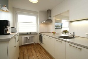 A modern kitchen with white cabinets, a built-in oven, a stainless steel sink, and a window providing natural light. - "Sandplaat" in Uns to Hus (Juist)