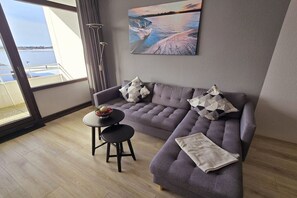 Ein modernes Wohnzimmer mit einem grauen Sofa, einem kleinen runden Tisch mit einer Obstschale und einem Gemälde einer Küstenlandschaft.