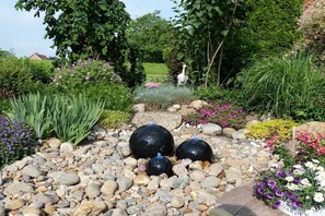 Ein Garten mit verschiedenen Pflanzen, einer Vogel-Skulptur und drei kugelförmigen Objekten auf einem Kiesbett.
