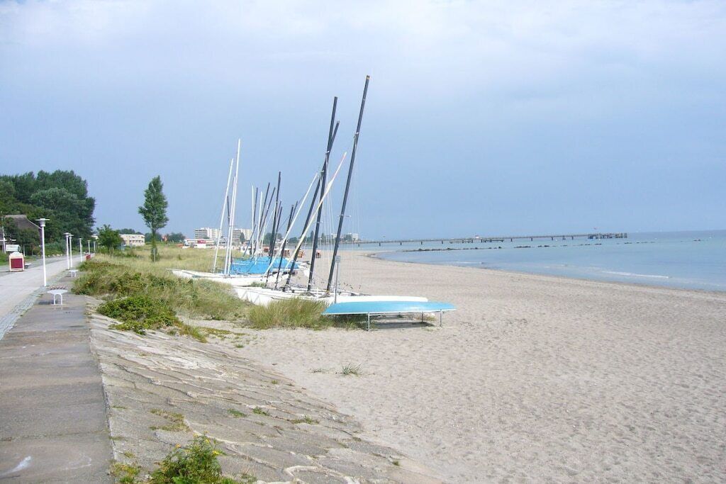 Una playa con una hilera de veleros amarrados a la orilla, un paseo marítimo de hormigón y un muelle que se adentra en el agua.