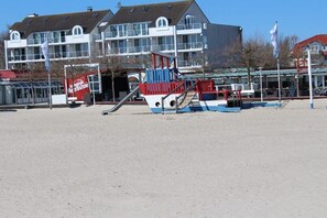 Een speeltuin aan het strand met een glijbaan en klimconstructies, een gebouw met meerdere ramen, en een vlaggenmast.