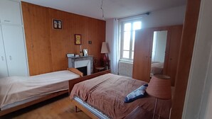 Une chambre avec deux lits simples, une armoire en bois, un miroir, une cheminée et une fenêtre avec des rideaux.