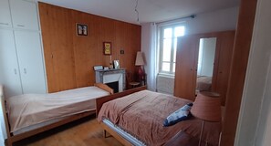 A bedroom with two single beds, a wooden wardrobe, a mirror, a fireplace, and a window with curtains. - Village House with Courtyard near Issoire (Perrier)