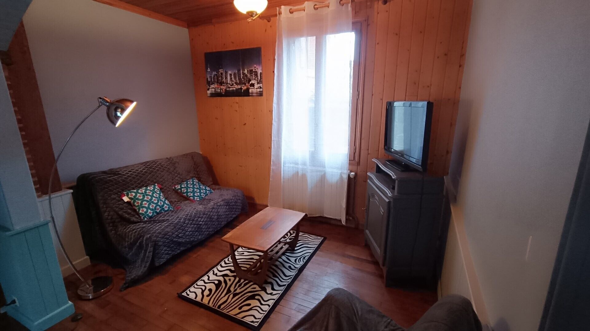 Ein gemütliches Wohnzimmer mit einer Couch, einem Flachbildfernseher, einer Lampe und einem Stadtbild-Poster an der Wand.