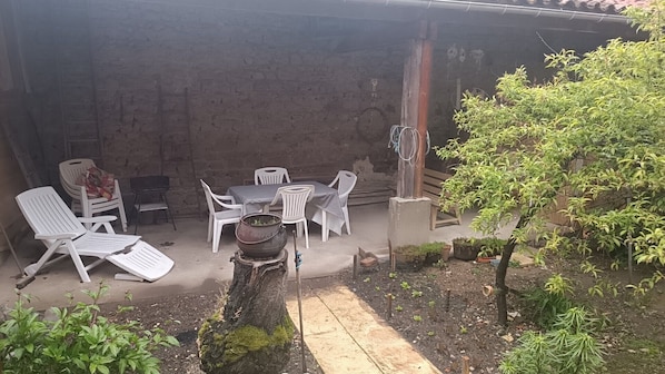 A covered outdoor seating area with white plastic chairs and a table, surrounded by plants and a tree.