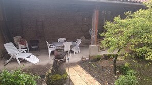 A covered outdoor seating area with white plastic chairs and a table, surrounded by plants and a tree. - Village House with Courtyard near Issoire (Perrier)