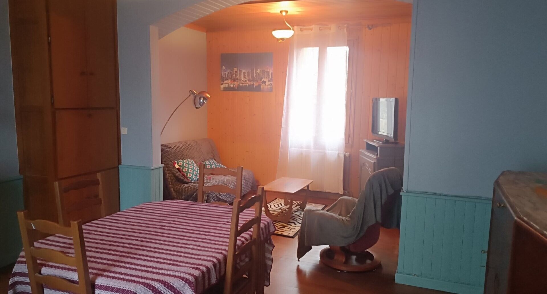 Ein kleines Zimmer mit einem Bett, einem Sessel, einem kleinen Tisch, einem Fernseher und einem Bild an der Wand.