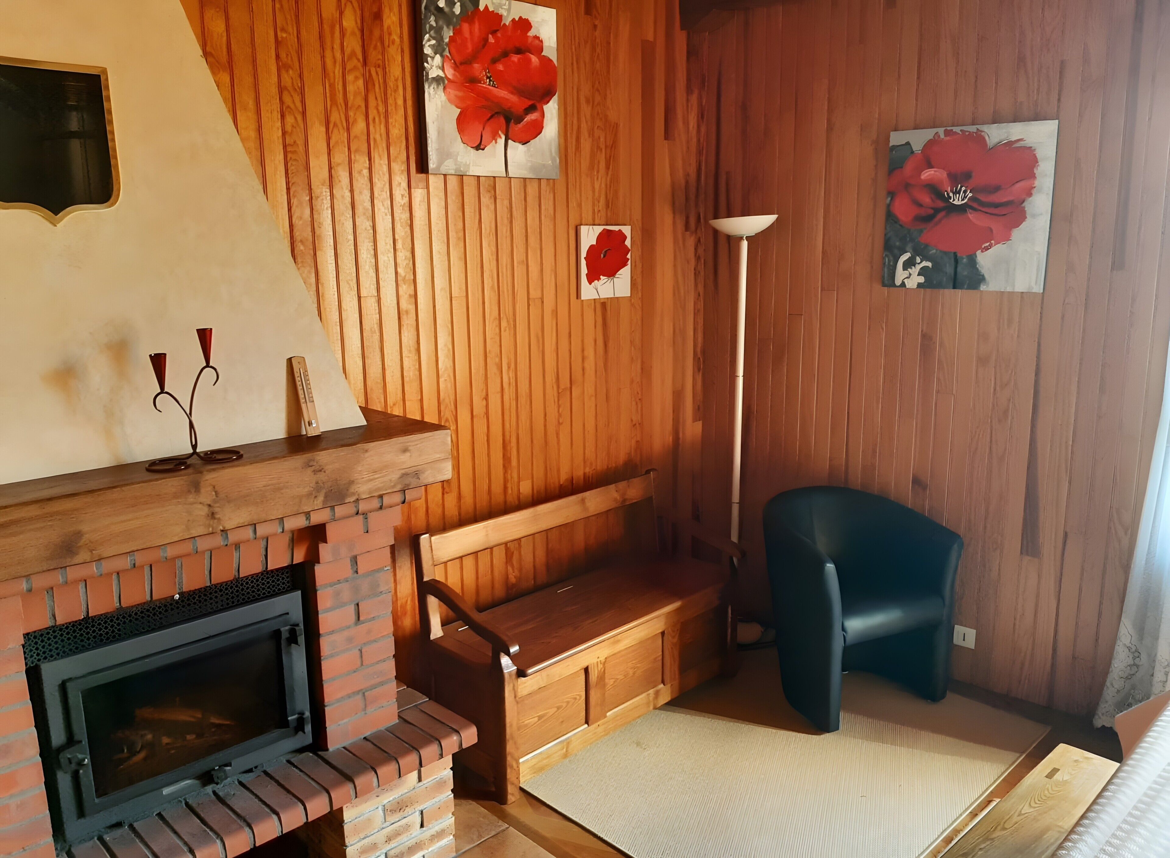 A cozy room with a brick fireplace, wooden bench, and two red flower paintings on the wall.