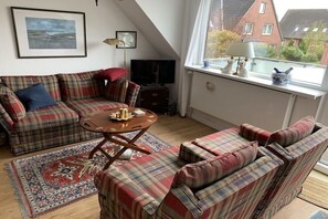 A living room with a sofa set, a coffee table, a rug, a television, and a window with a view of buildings. - Apartment building "Albatros" (St. Peter-Ording)