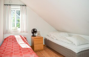 Een kleine, goed verlichte kamer met een schuine kap, een bed met wit beddengoed, een nachtkastje met een ventilator en een raam met gordijnen.