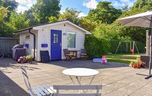 Een laag, enkel verhaal tellend gebouw met een blauwe deur en ramen, een terras met een ronde tafel en stoelen, en een schommelset op de achtergrond.