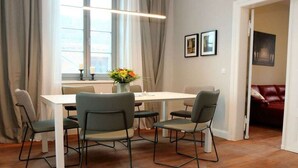 Uma sala de jantar com uma mesa branca e quatro cadeiras, um vaso com flores e um sofá vermelho ao fundo.