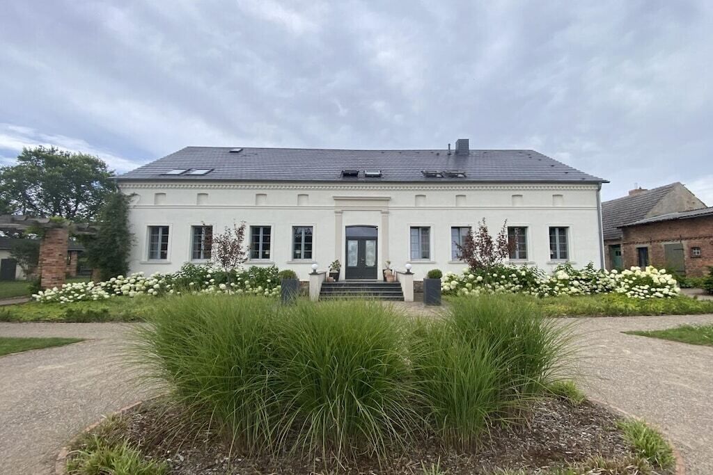 En toetages bygning med en symmetrisk facade, en central indgang med trapper, og en have med høje græstyper og hvide blomster foran.