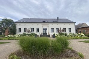 Ein zweigeschossiges Gebäude mit einer symmetrischen Fassade, einem zentralen Eingang mit Stufen und einem Vorgarten mit hohem Gras und weißen Blüten.