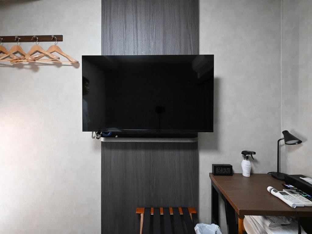 Een kamer met een tv aan de muur, een houten bank, een bureau met een lamp en een wandklok.