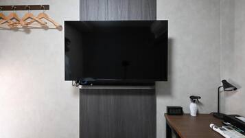 Een kamer met een tv aan de muur, een houten bank, een bureau met een lamp en een wandklok.