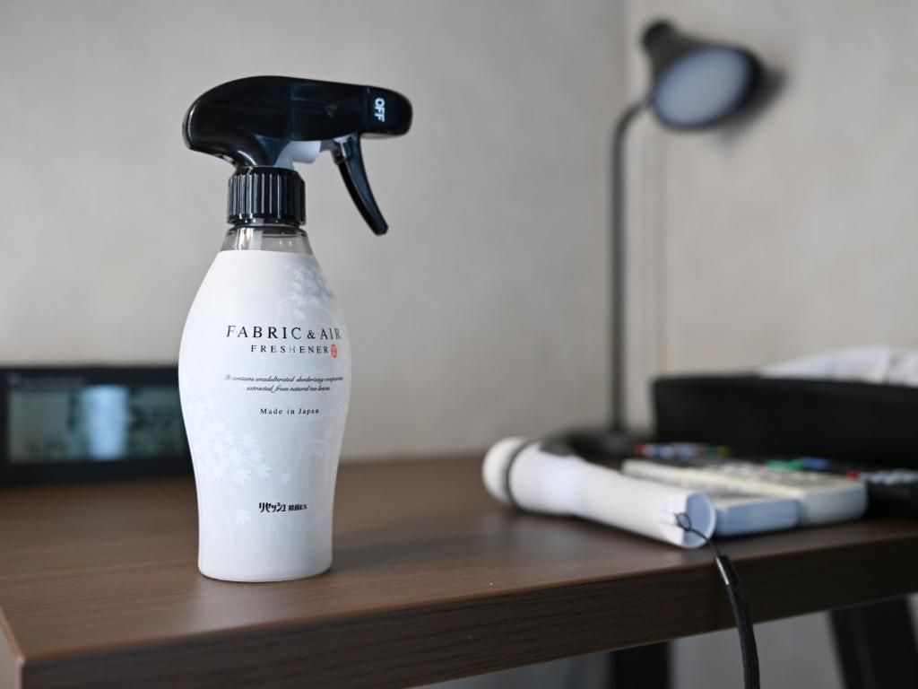 Un atomizador etiquetado como 'FABRIC & AIR FRESHENER' sobre una superficie de madera, con una lámpara y un control remoto al fondo.