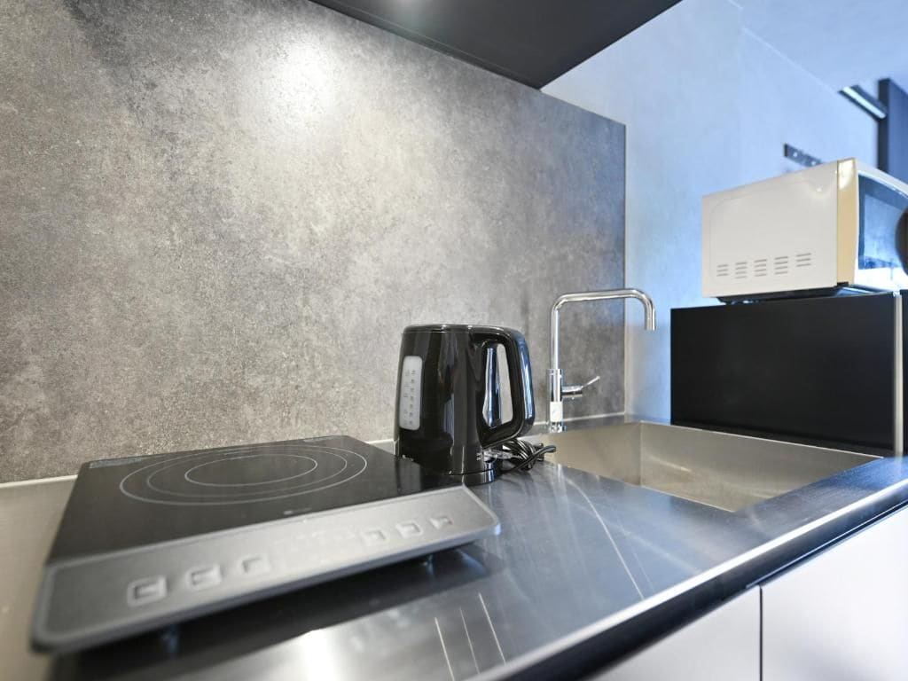 Una cocina con una placa vitrocerámica moderna, un hervidor y un microondas sobre una encimera de acero inoxidable.