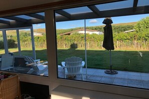 Habitació amb una finestra gran que dona a un jardí, una cadira blanca i una taula amb un ordinador portàtil.