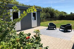A small building with a door and windows, a paved patio with two chairs, and a garden with green foliage and red berries. - Holiday home with 1 bedroom (Julianadorp)